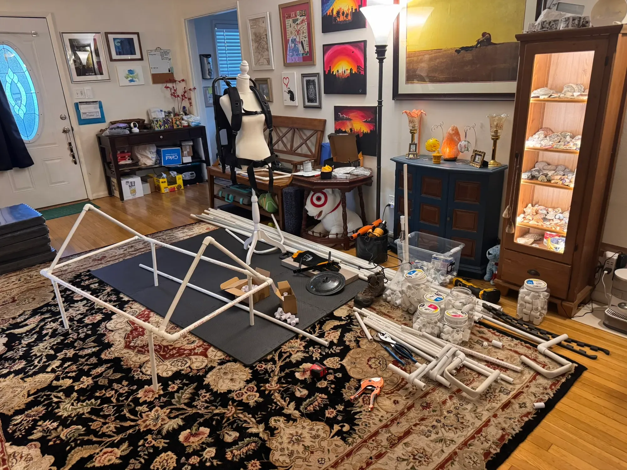 The living room as staging area: dry-fit PVC sections, a dress form wearing the black pack harness, and jars of fittings everywhere