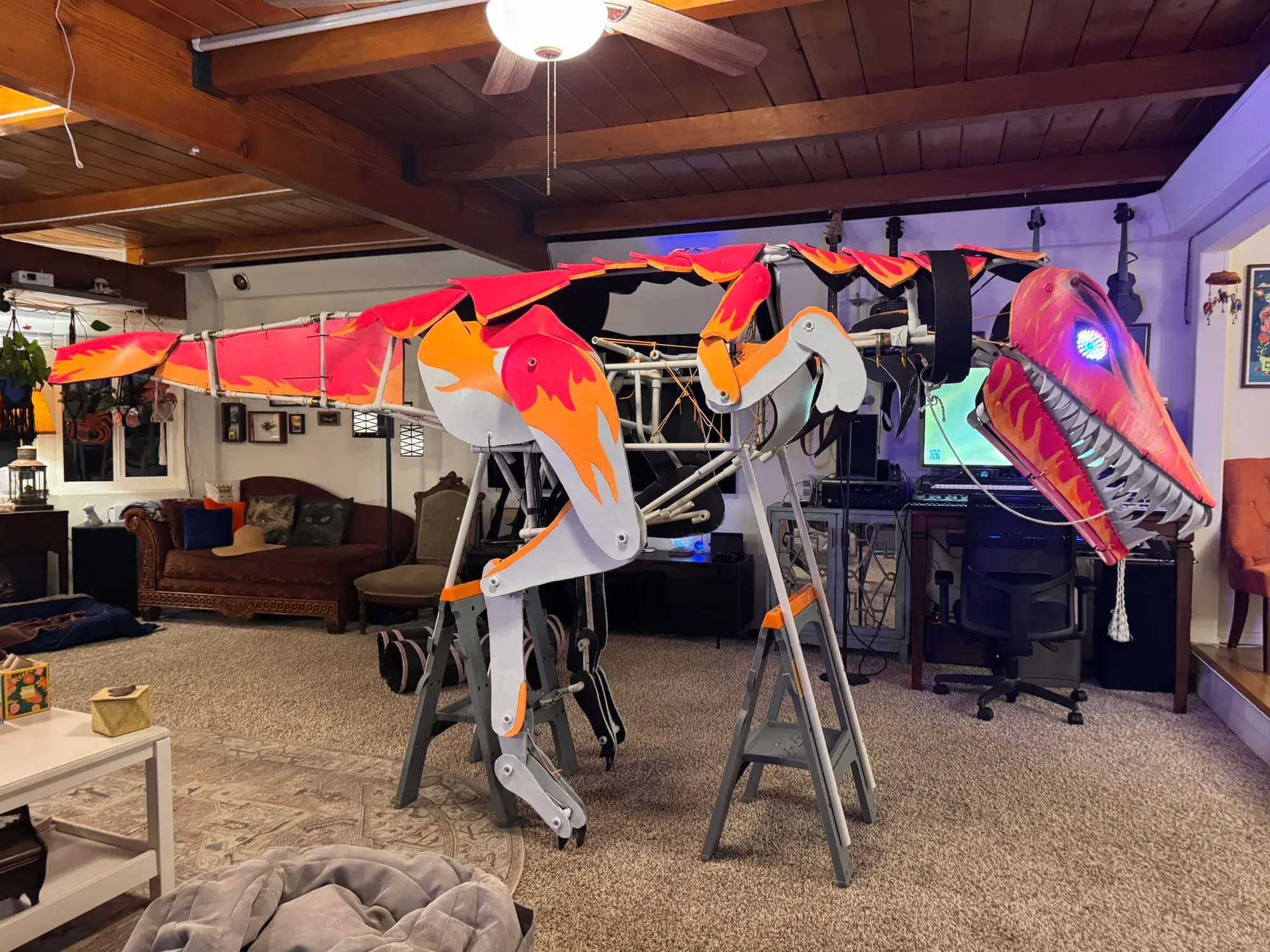 The finished raptor on sawhorses in the living room: flame-striped pink and orange body plates, gray and orange legs, a toothy head, and one glowing blue-violet LED eye